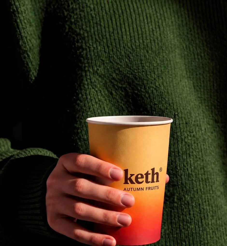 A hand holding an orange Keth Autumn Fruits cup against a green knit sweater